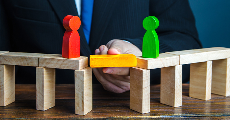 TeamPerform_3 • Roche Leadership A person in a suit bridges a gap between two wooden platforms using a yellow block, connecting red and green figures representing teamwork or mediation.