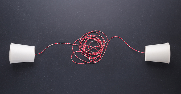 TeamPerform_6 • Roche Leadership Two paper cups connected by a red and white tangled string on a dark surface, representing complex or unclear team communication.