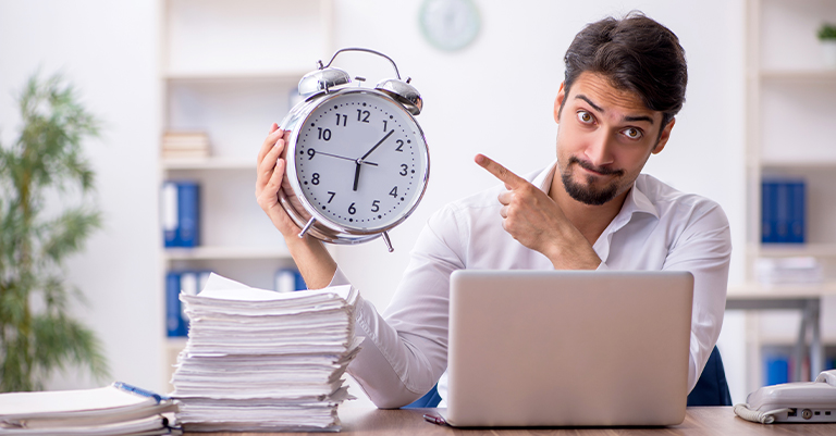 TimeMgmt_1 • Roche Leadership Man pointing at a large clock while sitting at a desk with paperwork and a laptop.